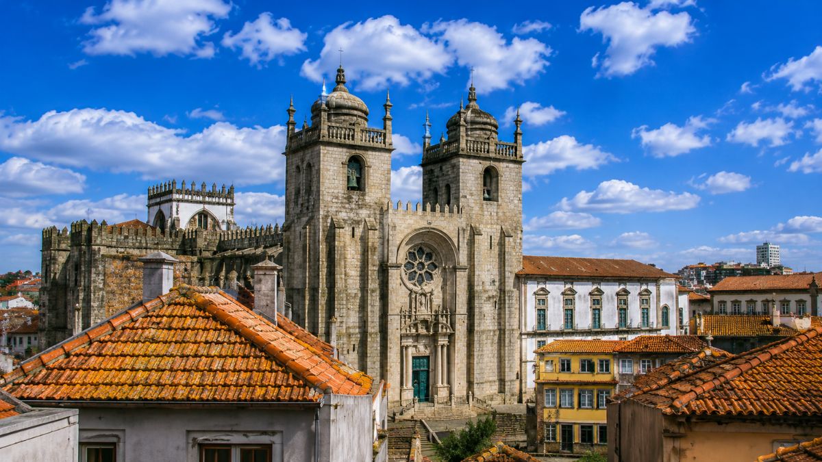 Vista panorâmica urbana dos telhados de cerâmica laranja do Porto, revelando a imponente fachada de granito da Catedral da Sé com suas duas torres sineiras e rosácea central sob um céu azul com nuvens.