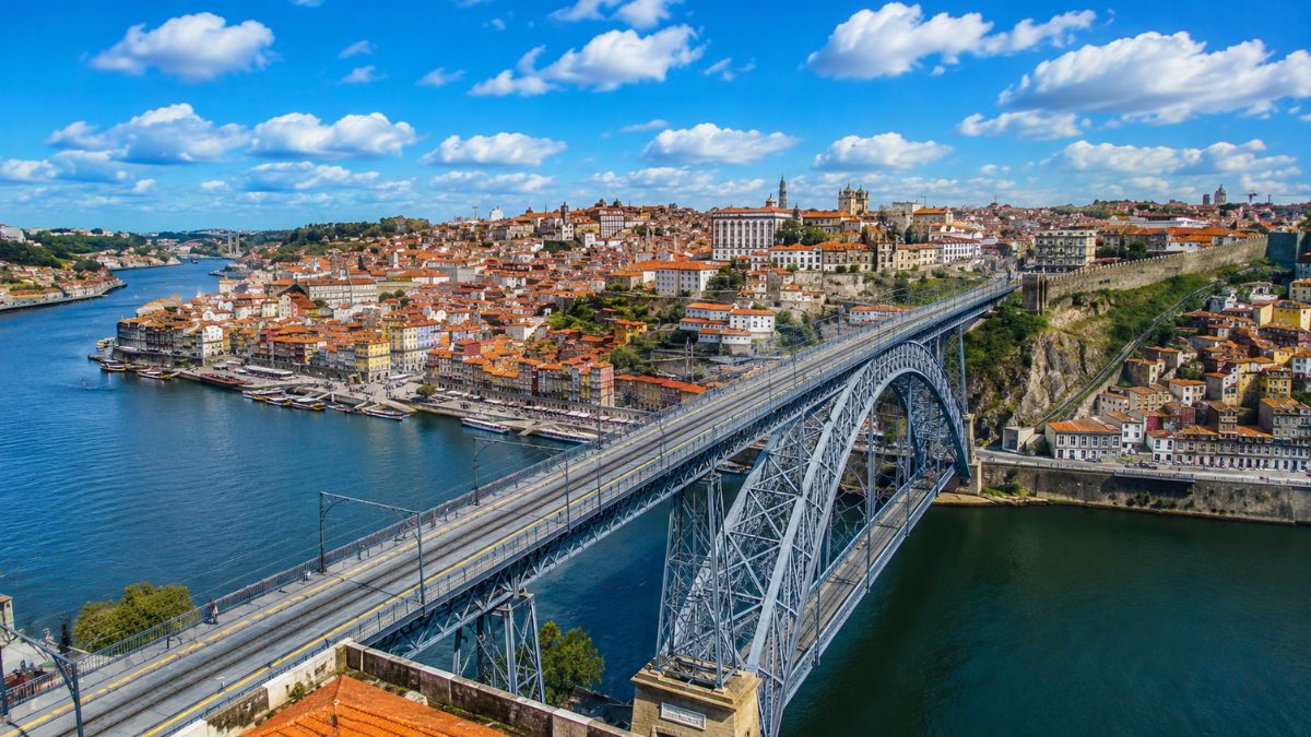 Fotografia aérea da Ponte Dom Luís I sobre o Rio Douro, destacando a arquitetura metálica, as águas azuis, o casario colorido da Ribeira e o céu ensolarado na cidade do Porto.