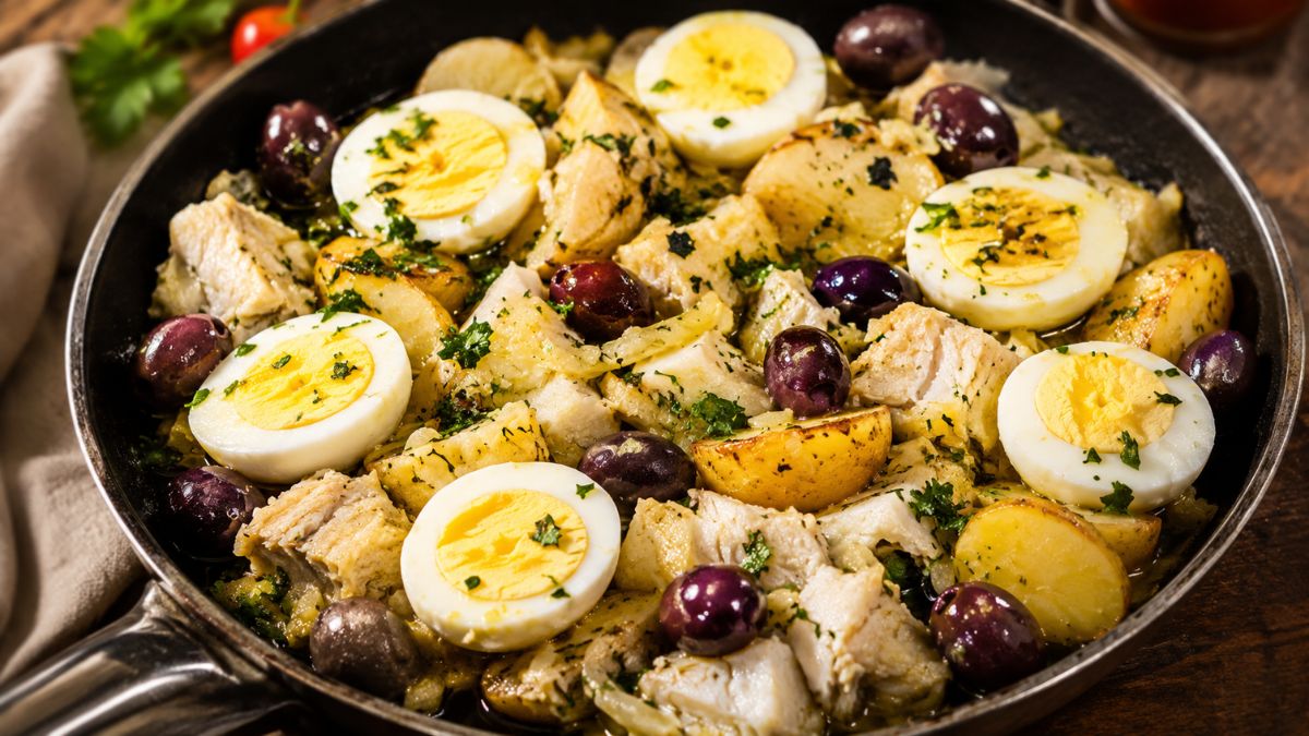 Fotografia em plano próximo de uma panela de ferro preta contendo o prato português Bacalhau à Gomes de Sá. A imagem mostra lascas de bacalhau misturadas com batatas douradas, azeitonas pretas inteiras e cebolas finas. Por cima, há várias rodelas de ovos cozidos com a gema aparente e tudo está salpicado com salsa fresca picada em um ambiente rústico com luz natural.