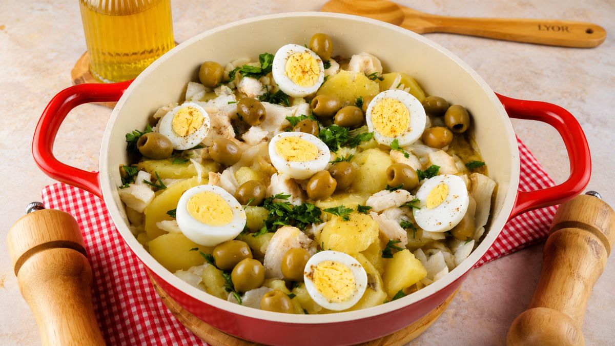 Panela vermelha com bacalhoada tradicional portuguesa misturada com batatas cozidas, decorada com azeitonas verdes inteiras, ovos cozidos cortados ao meio e salsa fresca picada em cima. Ao fundo, garrafa de azeite e utensílios de madeira.