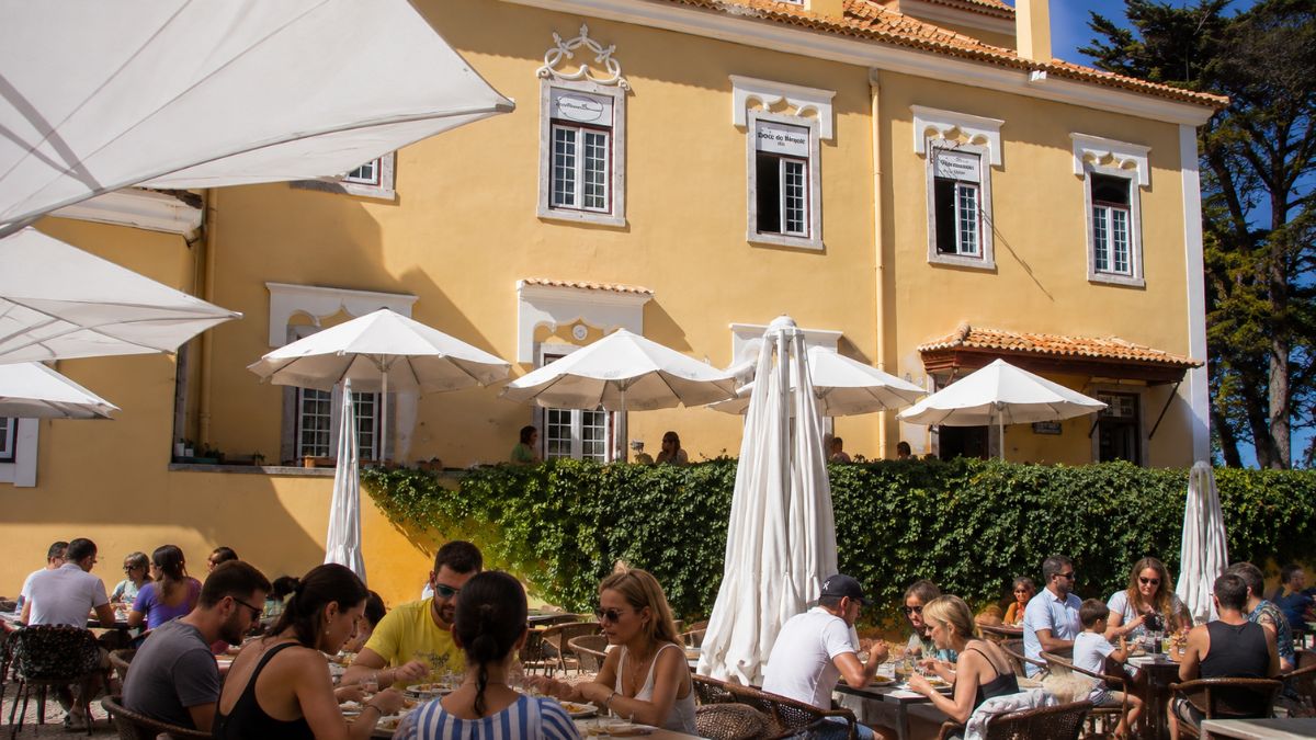 Pessoas almoçando na esplanada com guarda-sóis brancos em frente ao palacete amarelo da Casa da Guia em Cascais.