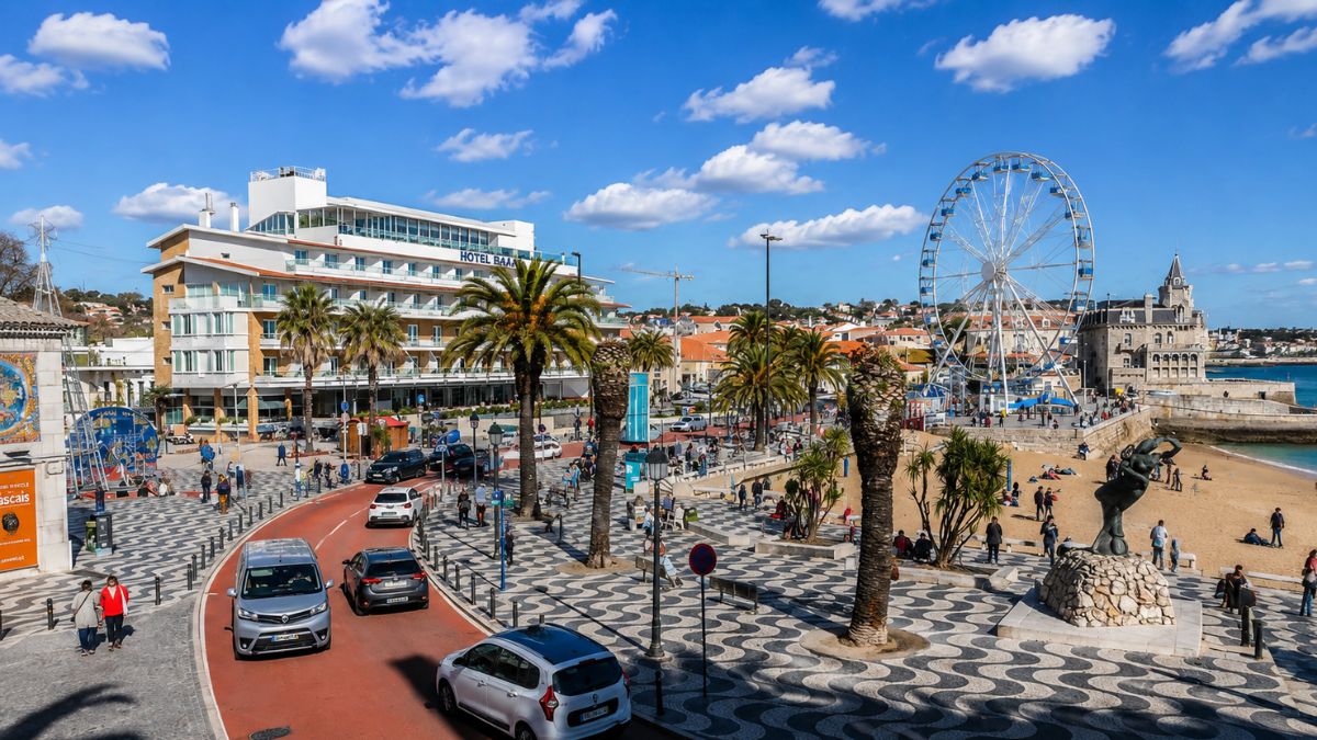 Fotografia da orla marítima da vila de Cascais com calçada portuguesa, roda-gigante, praia da baía e palacete histórico ao fundo.