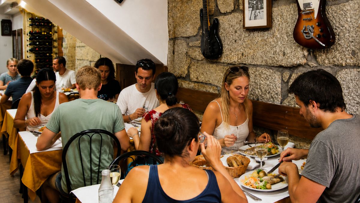 Ambiente interno de restaurante tradicional no Porto com pessoas comendo pratos portugueses em mesas com toalha branca perto de parede de pedra.