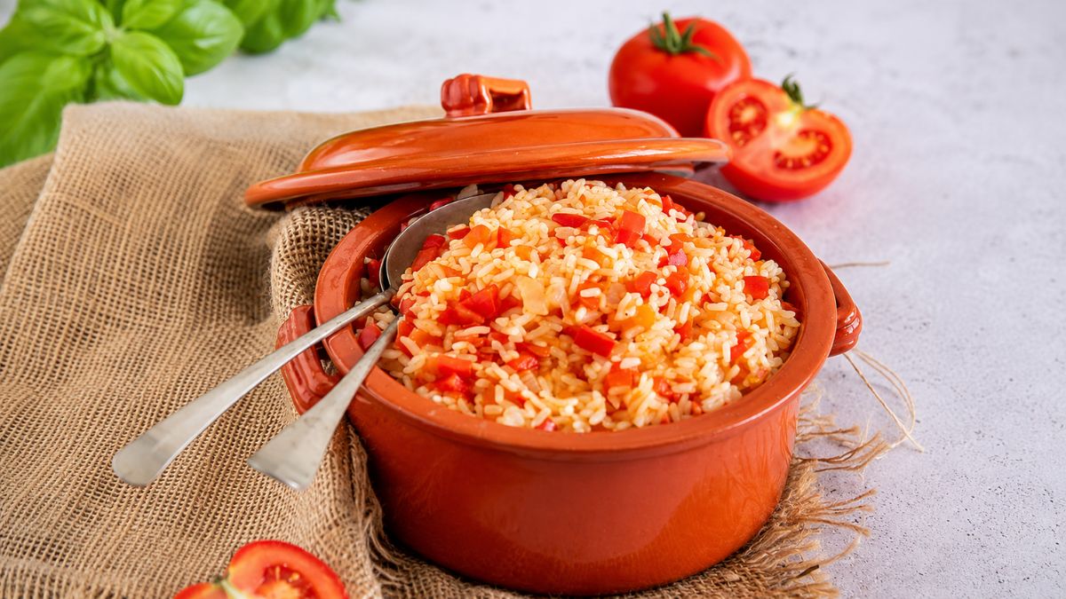 Panela de barro com arroz de tomate e pimentão vermelho, acompanhada por tomates frescos e manjericão sobre pano rústico.