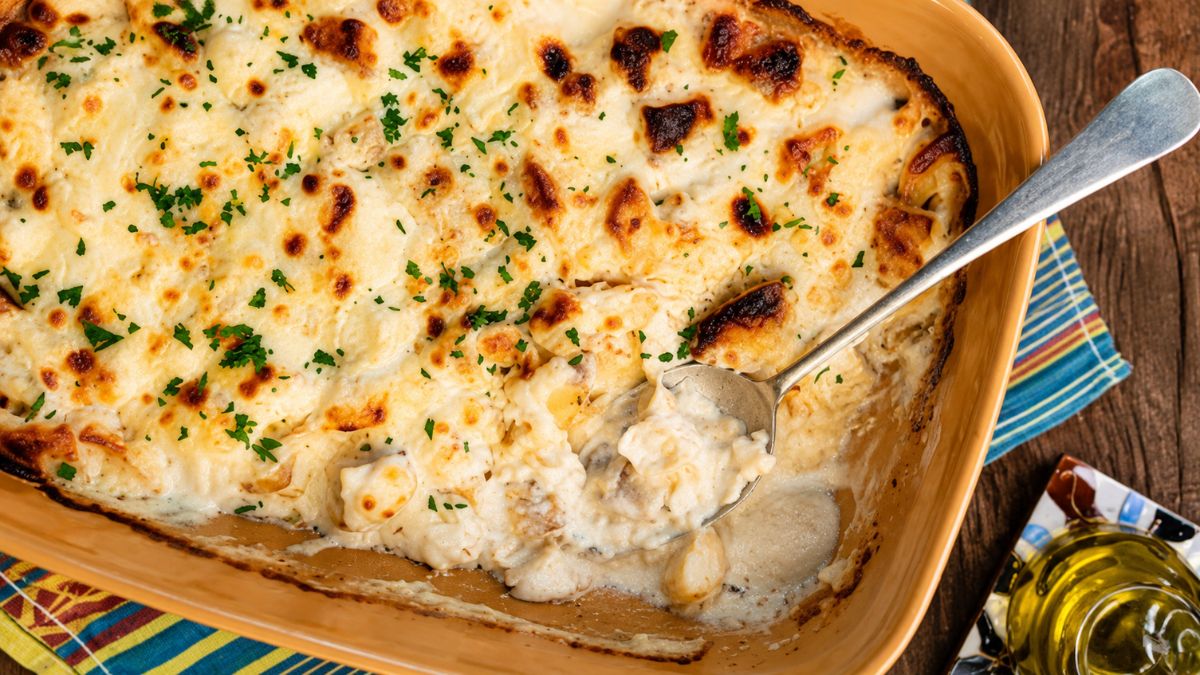 Travessa refratária amarela com bacalhau com natas gratinado dourado, salpicado de salsa picada e servido com colher de metal.