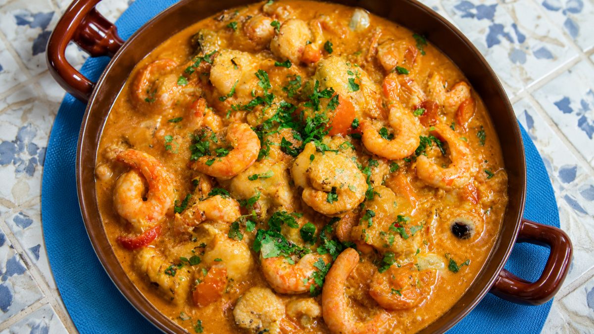 Travessa de barro com ensopado de camarões grandes e mariscos em molho cremoso alaranjado, polvilhado com salsa fresca picada.
