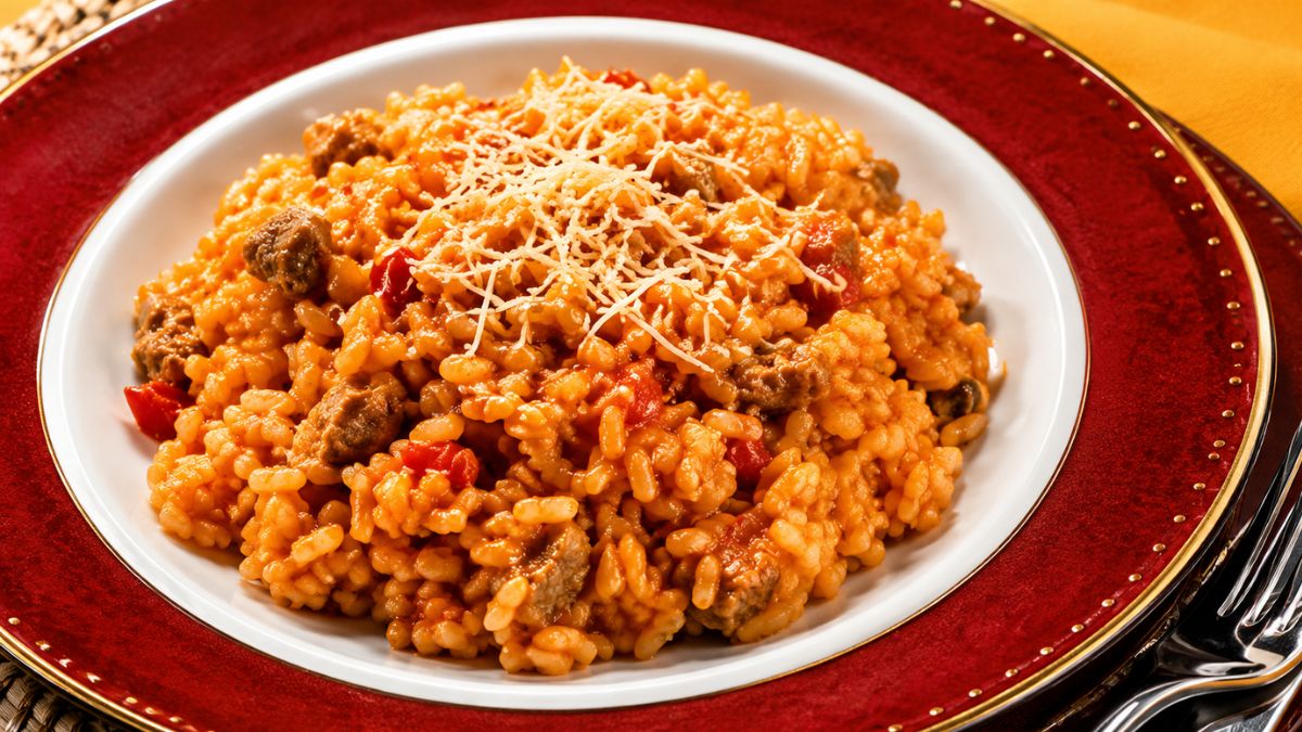 Prato fundo vermelho com risoto cremoso de tomate e pedaços de carne, finalizado com queijo ralado por cima.