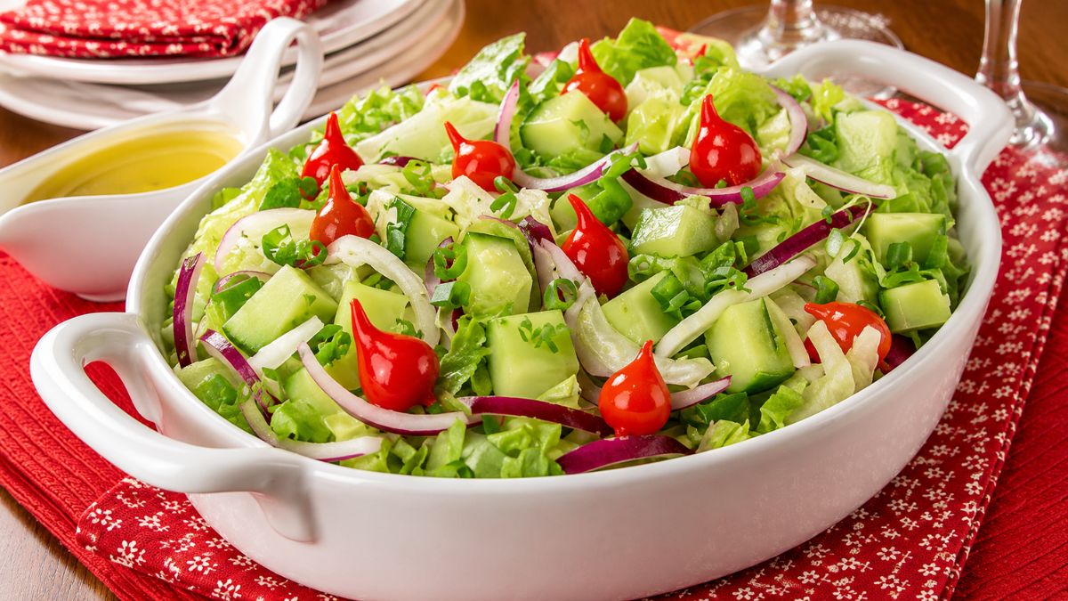 Travessa branca com salada de folhas verdes, pepino em cubos, cebola roxa e pimentas biquinho vermelhas com molheira ao fundo.