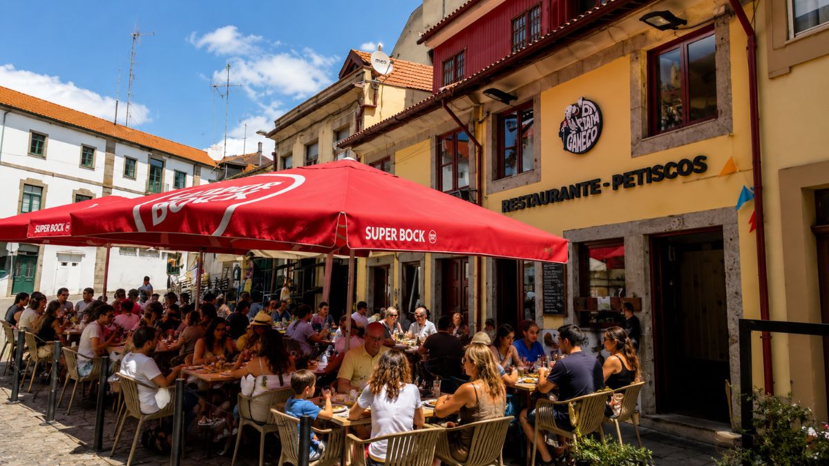 Esplanada movimentada de um restaurante tradicional em uma rua histórica do Porto com clientes almoçando sob guarda-sóis vermelhos.