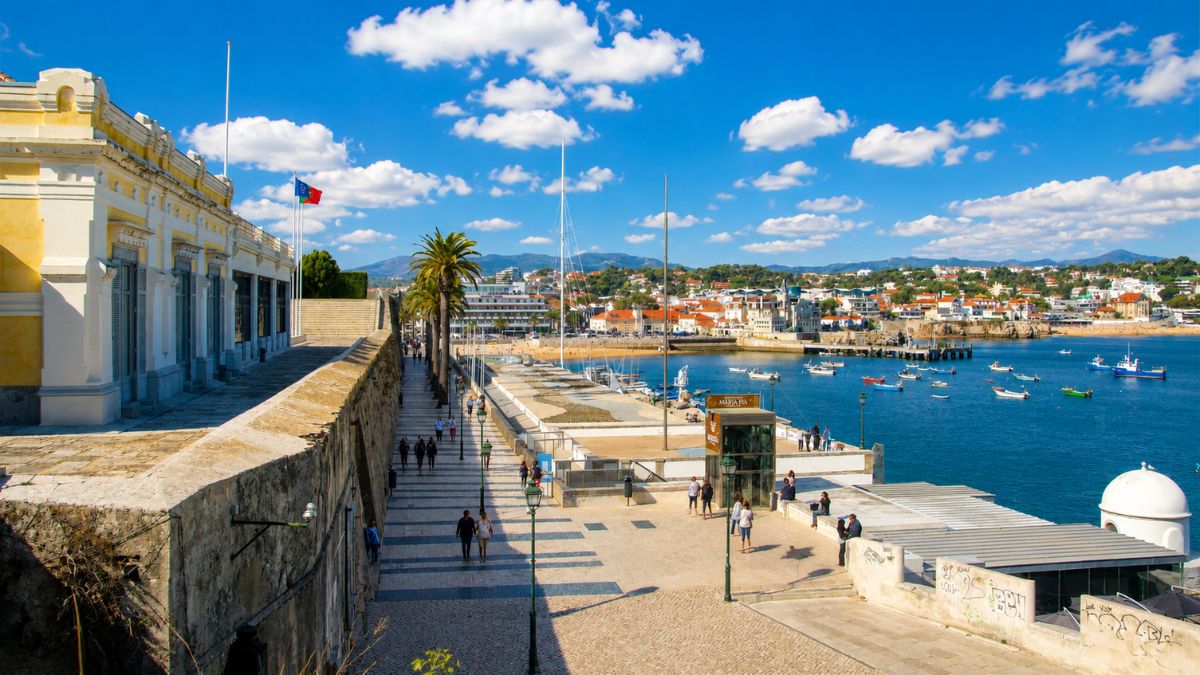 Fotografia da calçada litorânea e fortaleza de Cascais mostrando a baía com barcos de pesca e a Serra de Sintra no horizonte.