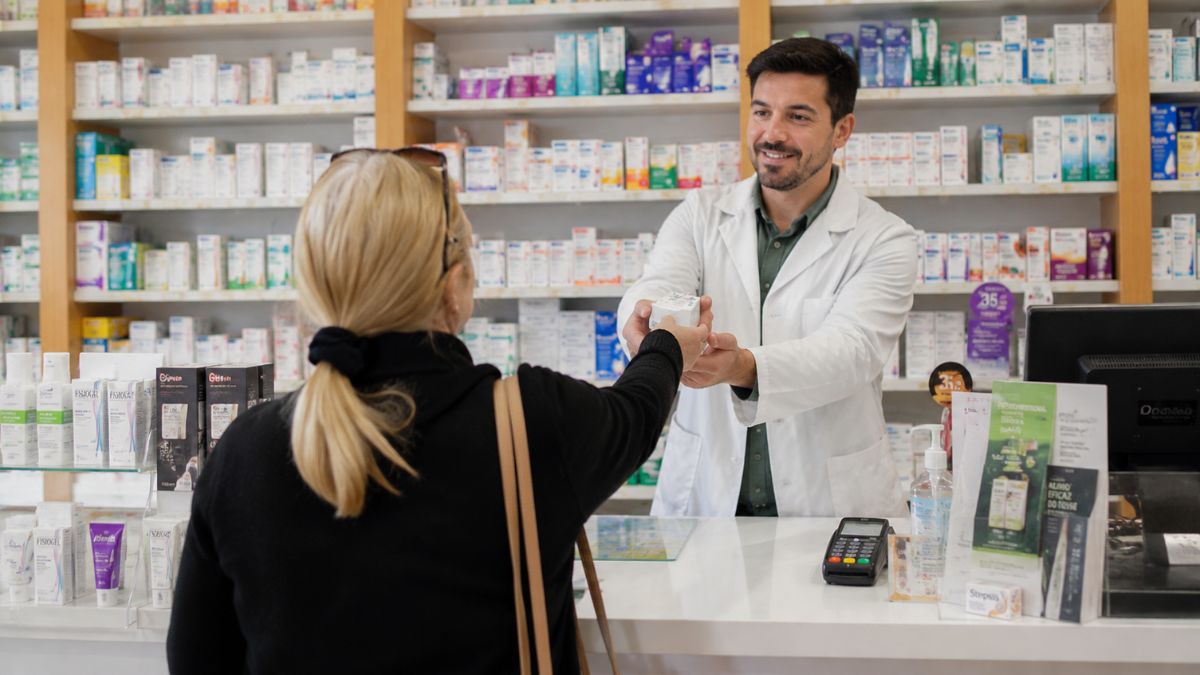 Farmacêutico com jaleco branco entregando caixa de remédio para uma cliente no balcão de uma farmácia em Arruda dos Vinhos.