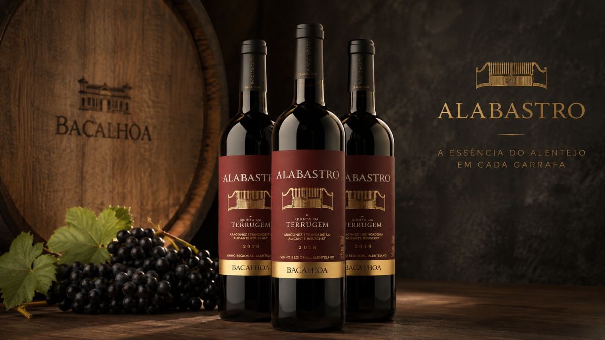 Três garrafas de vinho tinto Alabastro da Quinta da Terrugem colocadas sobre uma bancada de madeira, com o logótipo da Bacalhôa gravado no barril de carvalho ao fundo e a frase a essência do Alentejo em cada garrafa gravada em letras douradas à direita.