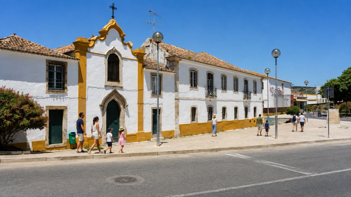 Moradores e pedestres caminhando pela calçada em frente a um prédio histórico branco e amarelo com capela em Arruda dos Vinhos.