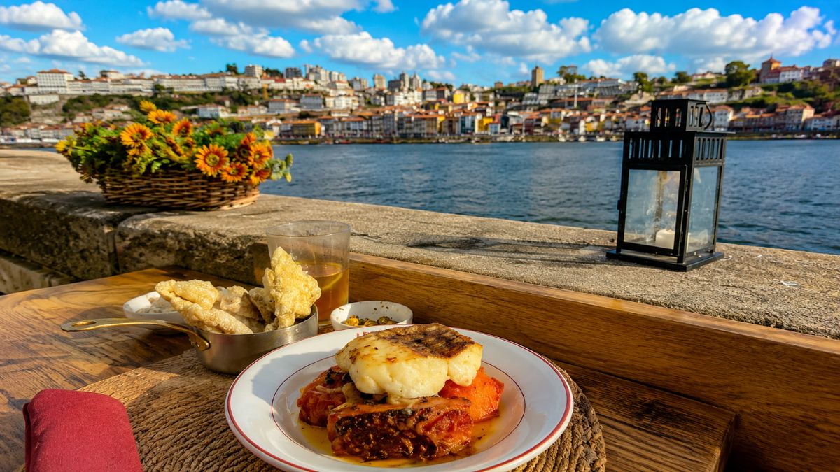 Prato tradicional de bacalhau servido em uma mesa à beira do rio Douro com a paisagem histórica da cidade do Porto ao fundo.