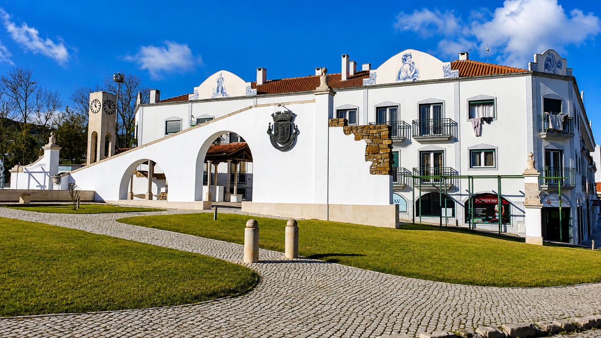 Praça pública arborizada com calçada portuguesa em pedras, arcos brancos decorativos, brasão e edifícios com arquitetura tradicional de Arruda dos Vinhos sob céu azul.