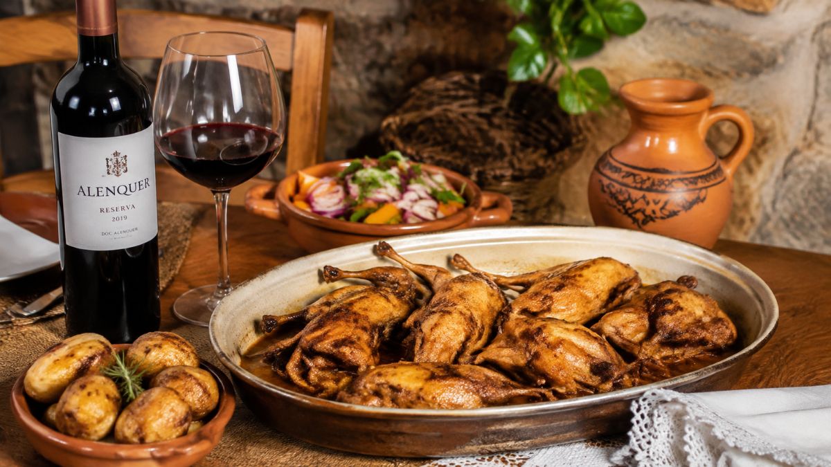 Travessa de barro com codornizes assadas no forno acompanhadas por batatas assadas, salada e uma garrafa de vinho tinto de Alenquer.