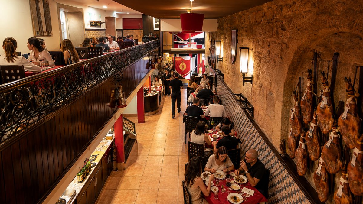 Interior do restaurante Abadia do Porto com clientes em mesas, paredes de pedra decoradas com presuntos curados e mezanino elegante.