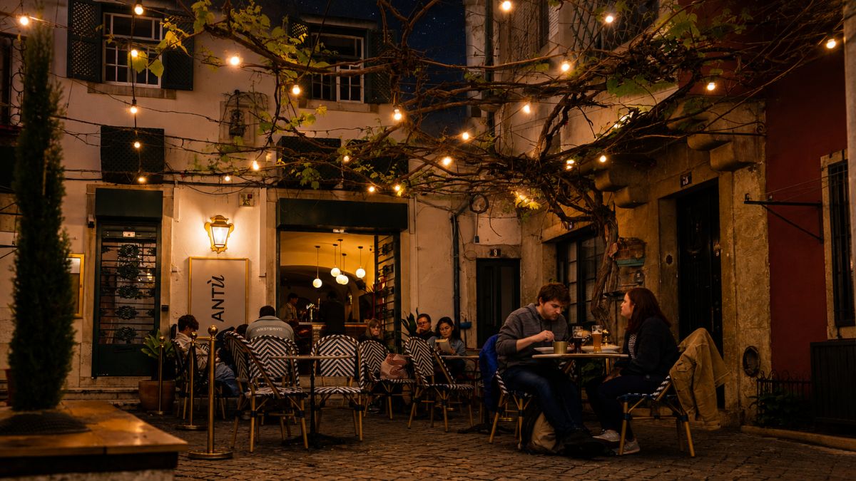 Pessoas jantando em uma esplanada charmosa de restaurante tradicional em Alfama à noite, iluminada por cordões de luzes sob parreiras e arquitetura típica lisboeta.