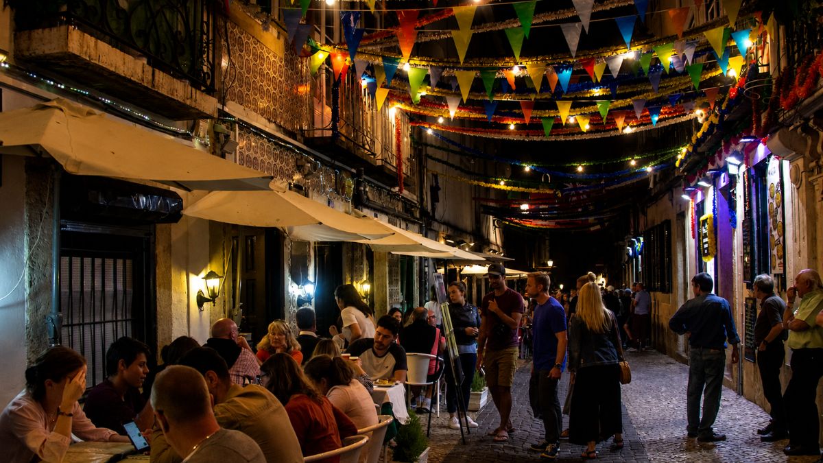 Pessoas caminhando e jantando em esplanadas ao ar livre em uma rua estreita de Lisboa decorada com bandeirolas coloridas e iluminação noturna.