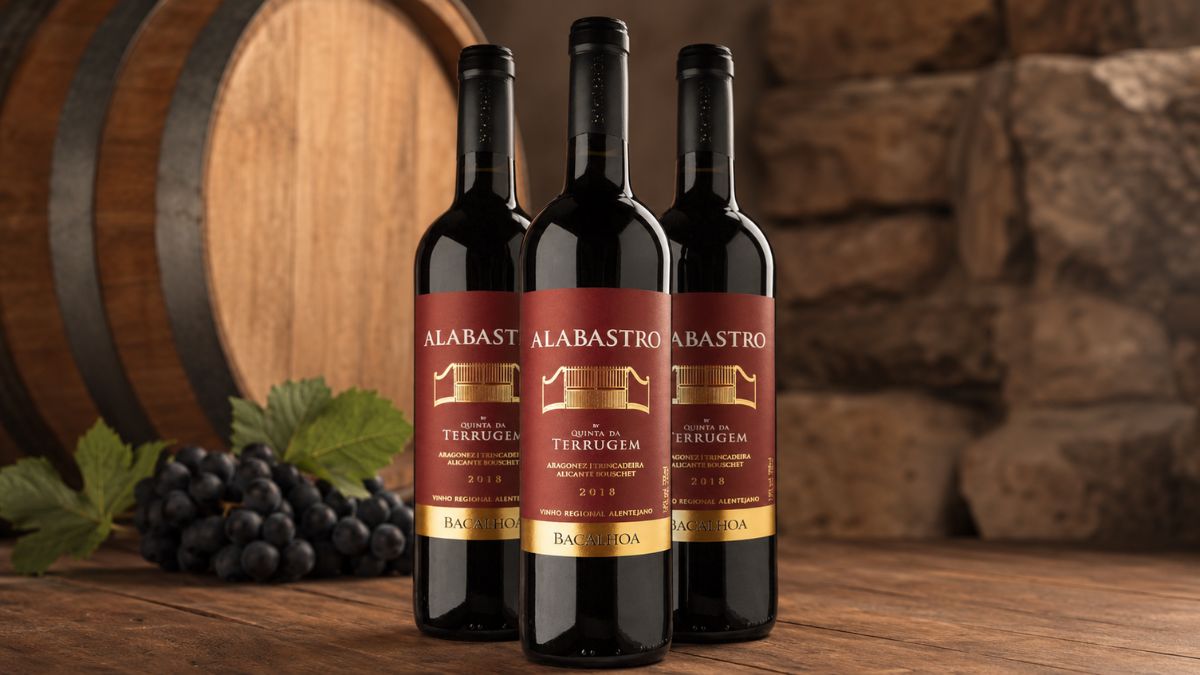 Imagem de estúdio foca três garrafas de vinho tinto Alabastro da Quinta da Terrugem em pé numa mesa de madeira rústica, com uvas roxas frescas e uma folha de videira à esquerda e um barril de carvalho e parede de pedra no fundo.