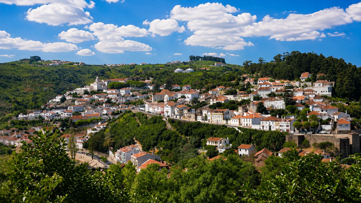 Vista panorâmica da encosta verdejante com o casario branco de Alenquer e as colinas da Serra do Montejunto sob céu azul.