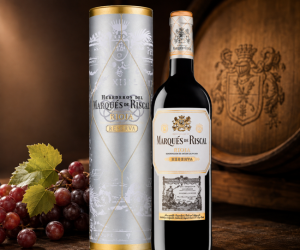 Garrafa de vinho tinto Marqués de Riscal Reserva Rioja ao lado do seu estojo metálico prata, com uvas vermelhas e barril de carvalho ao fundo.
