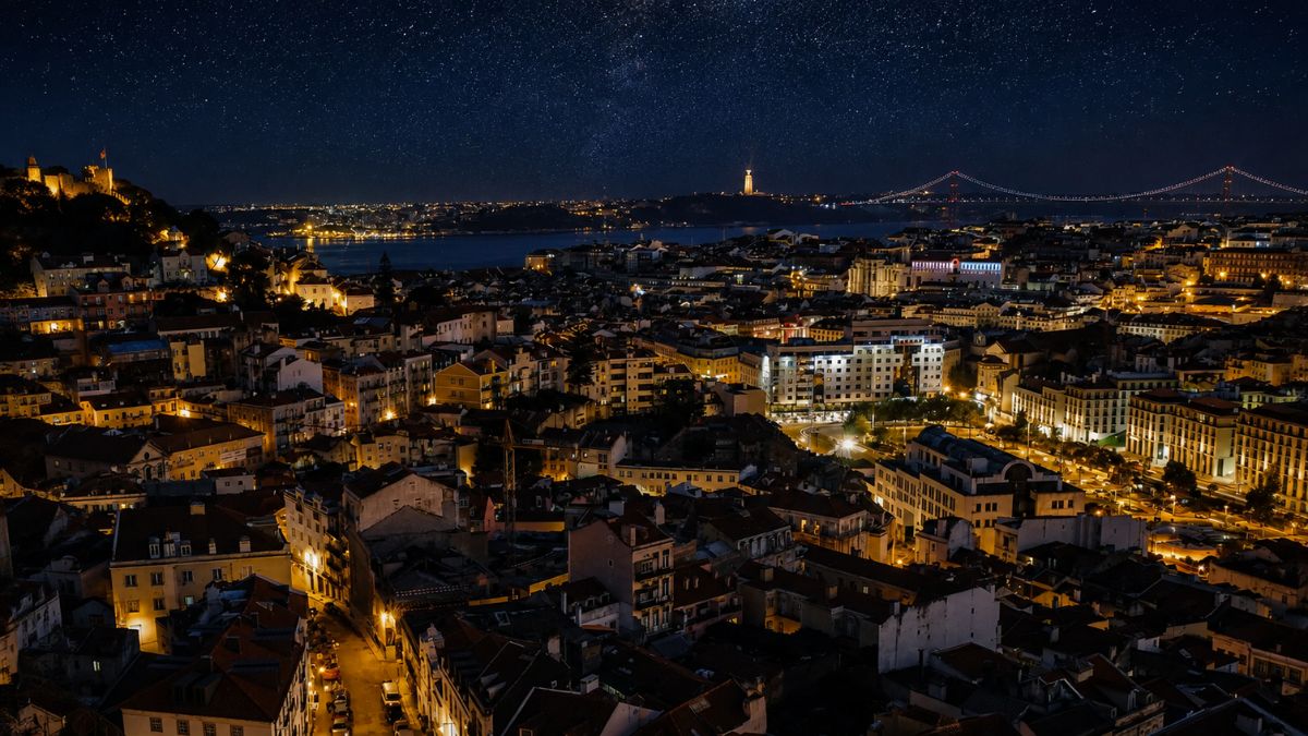 Fotografia noturna panorâmica da cidade de Lisboa iluminada, destacando o Castelo de São Jorge à esquerda, o Rio Tejo ao fundo, o Santuário do Cristo Rei e a Ponte 25 de Abril sob um céu estrelado.